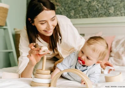 Mutter und Baby spielen mit einem Holzspielzeug auf einem Bett - In diesem Beitrag erfahren Sie, welches die sieben Tipps für gelungene Produktfotos von Babyartikeln sind.